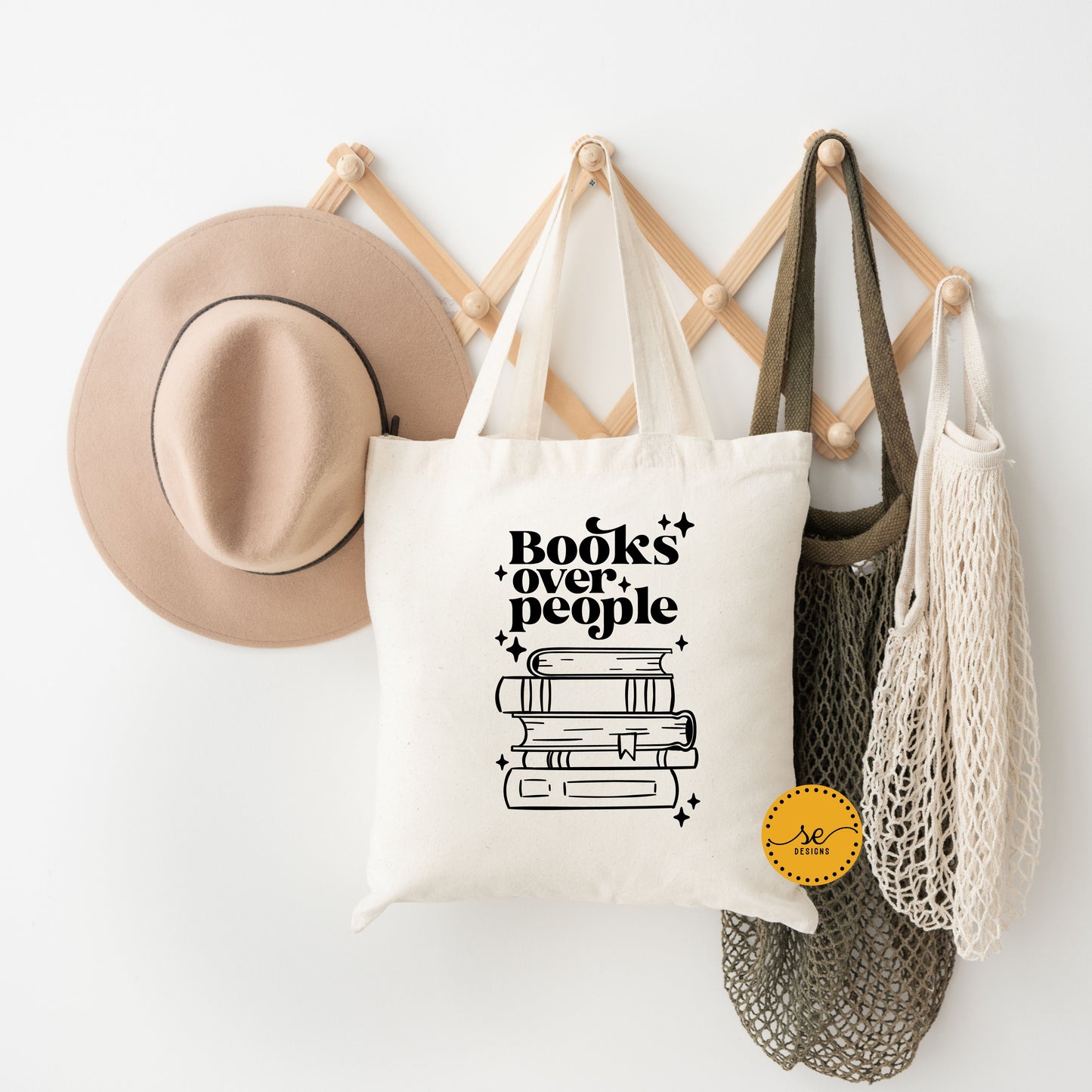"Neutral tote bag with 'Books over people' in bold black font above stacked books, accented with sparkles, hanging on a wooden peg rack with a hat and mesh bags."