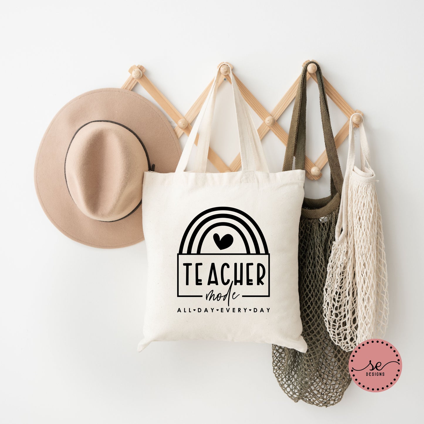 Teacher Mode All Day Every Day canvas tote bag featuring a minimalist rainbow design, hanging on a wooden rack next to neutral accessories